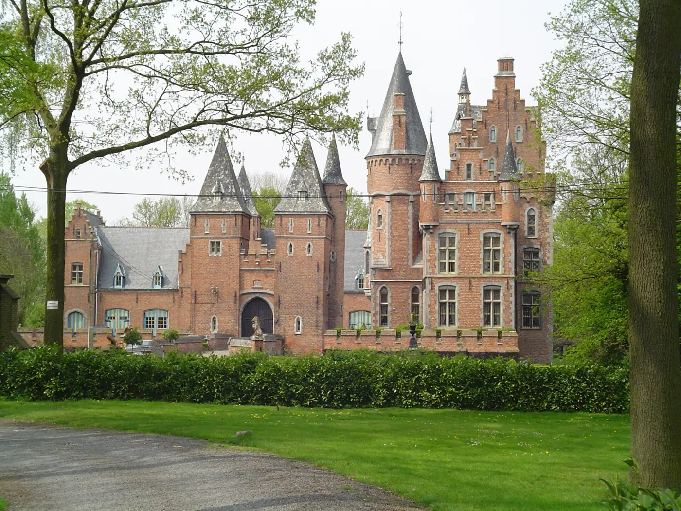 Het neogotische bakstenen Kasteel van Lovendegem met spitse torens en trapgevels, achter een haag en bomen.