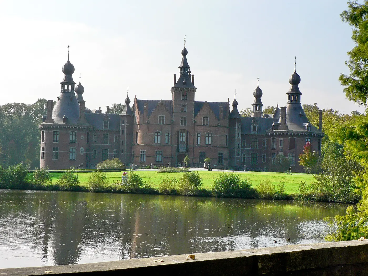 Kasteel Ooidonk met zijn torens weerspiegeld in de vijver, omringd door gazon en bomen.