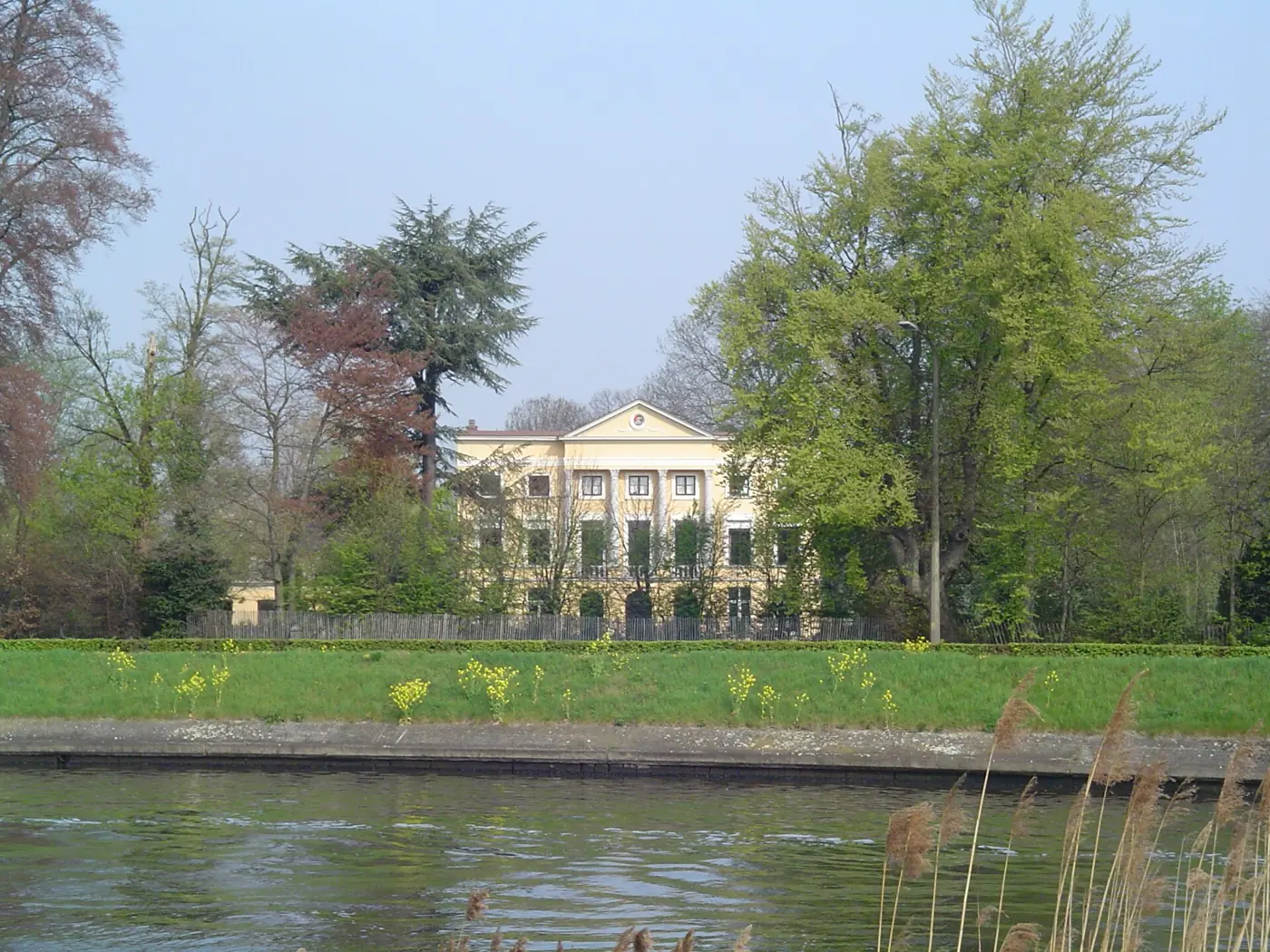 Het lichtgele neoclassicistische landhuis Oude Wal in Lovendegem, tussen bomen, gezien over een waterloop.